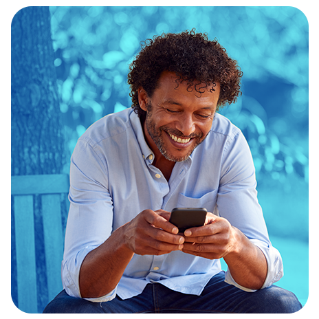 Homem sorrindo olhando o celular enquanto está sentado ao ar livre.