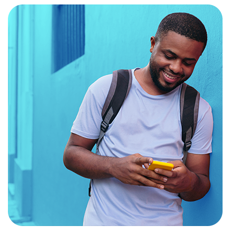 Homem com mochila sorrindo enquanto usa o celular encostado em uma parede.