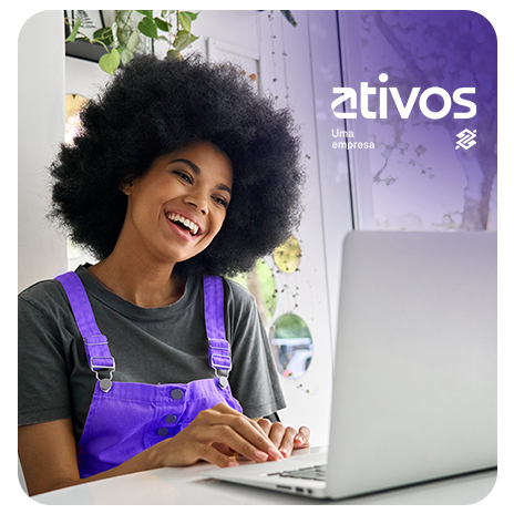 Mulher negra com cabelo black power sorri enquanto digita em um notebook. Ela veste uma camiseta cinza e um macacão roxo. Ao fundo, há uma janela com plantas penduradas e luz natural entrando no ambiente. No canto superior direito da imagem está o logotipo da Ativos acompanhado da frase "Uma empresa". O cenário transmite leveza, foco e positividade.