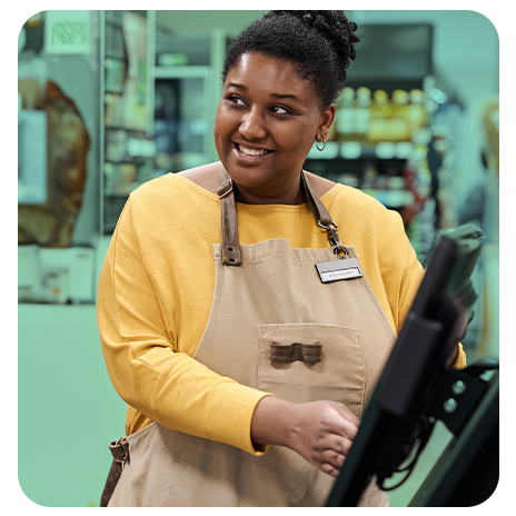 Mulher negra sorridente usando avental bege e blusa amarela, trabalhando em um caixa de supermercado ou comércio