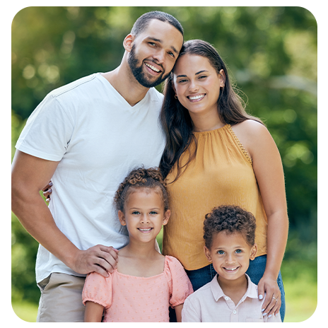 Família sorridente posando ao ar livre em um ambiente verde e ensolarado. 