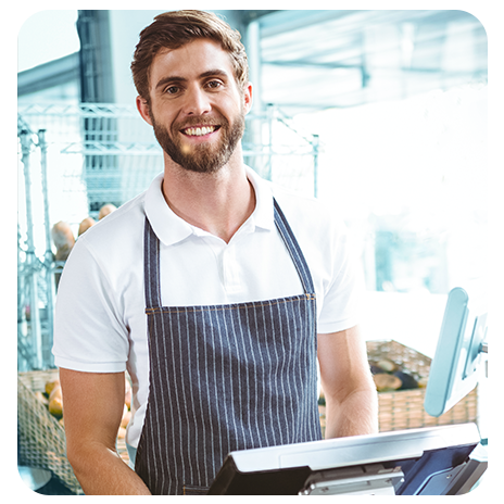 Homem jovem, branco, de barba e avental, sorrindo, atrás de um caixa de supermercado. 