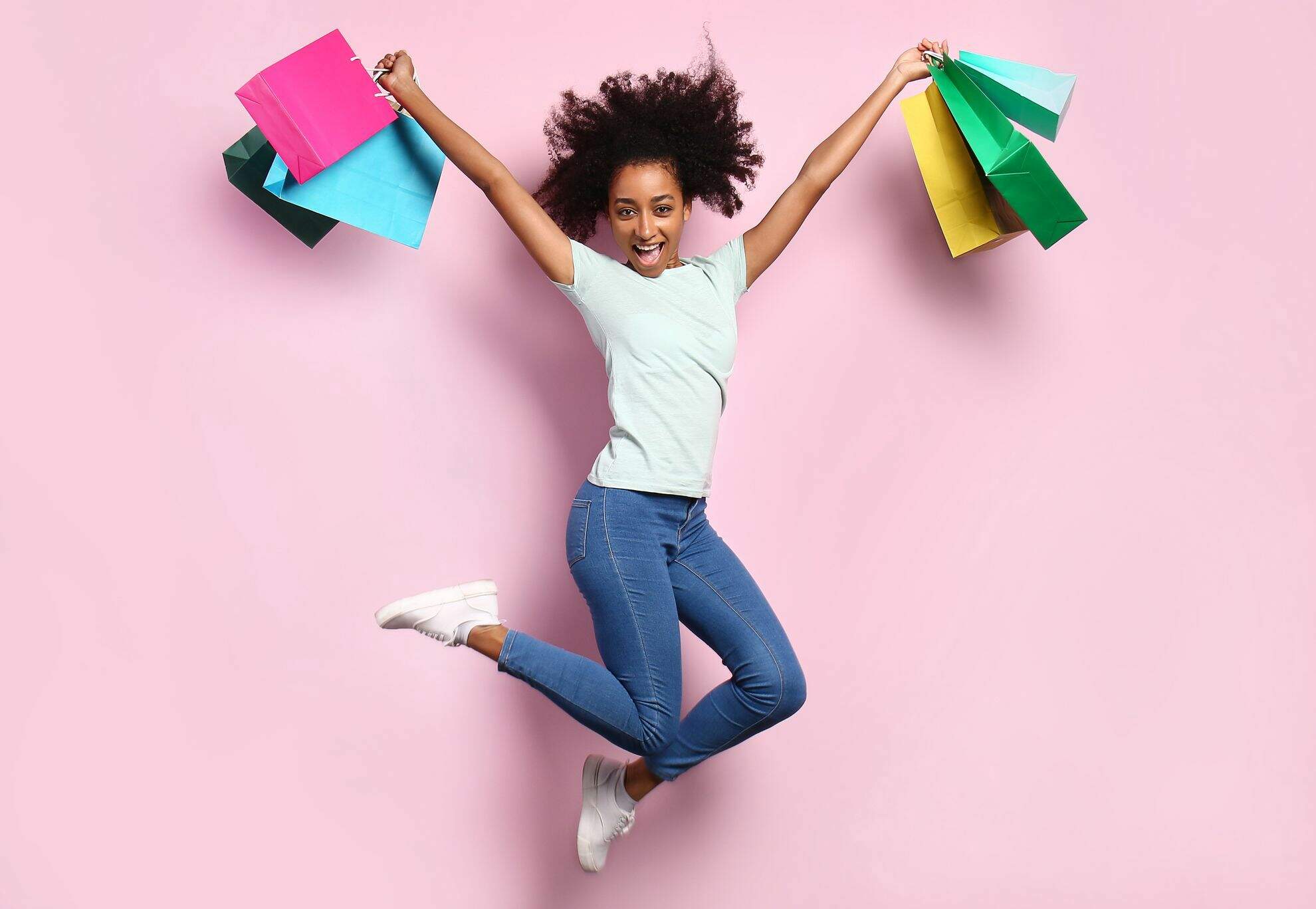 Jovem negra de camiseta branca, calça jeans e tenis, pulando feliz segurando várias sacolas coloridas de comepras nas duas mão em um fundo rosa.