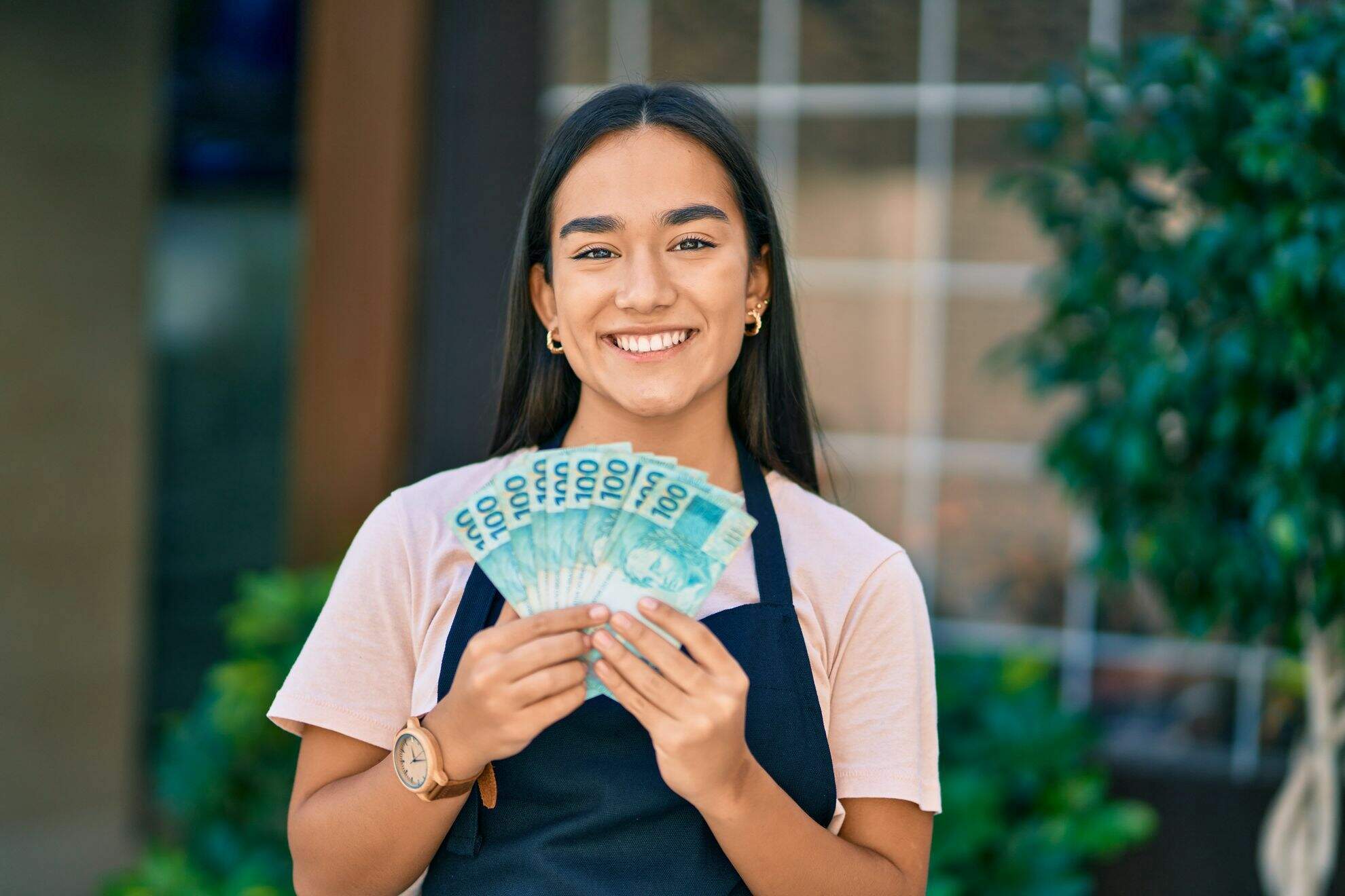 Mulher mestiça, sorrindo, segurando várias notas de cem reais com as duas mãos