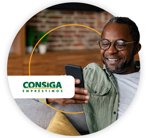 Homem negro de meia-idade, sorridente, usando óculos e camisa verde, sentado em um sofá enquanto olha para o celular. Ao fundo, ambiente doméstico com parede de tijolos e móveis de madeira. Sobre a imagem, há o logotipo da Consiga Empréstimos.