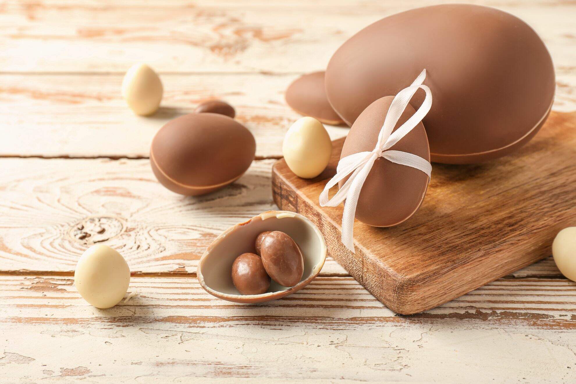 Bancada de madeira com um ovo grande de chocolate fechado, dois menores fechados, um deles com um laço branco, um menor aberto com ovinhos menores dentro, cercado por três ovos de galinha.
