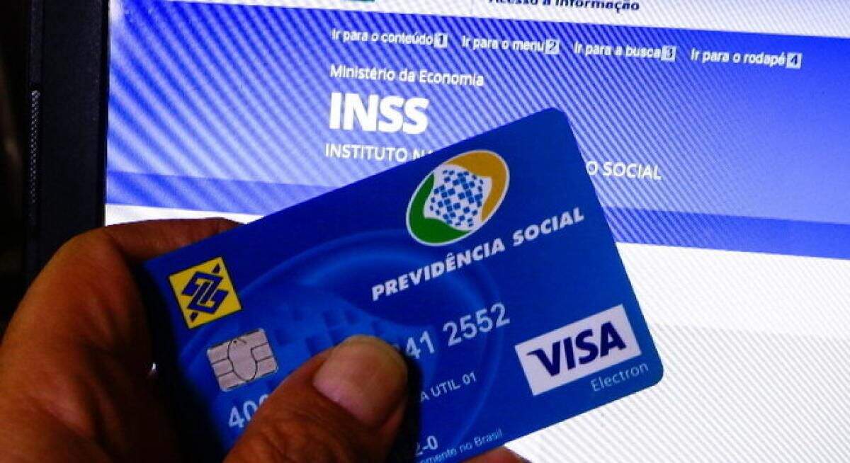 mão masculina segurando cartão do INSS em frente a tela de notebook aberta na tela do site do INSS.