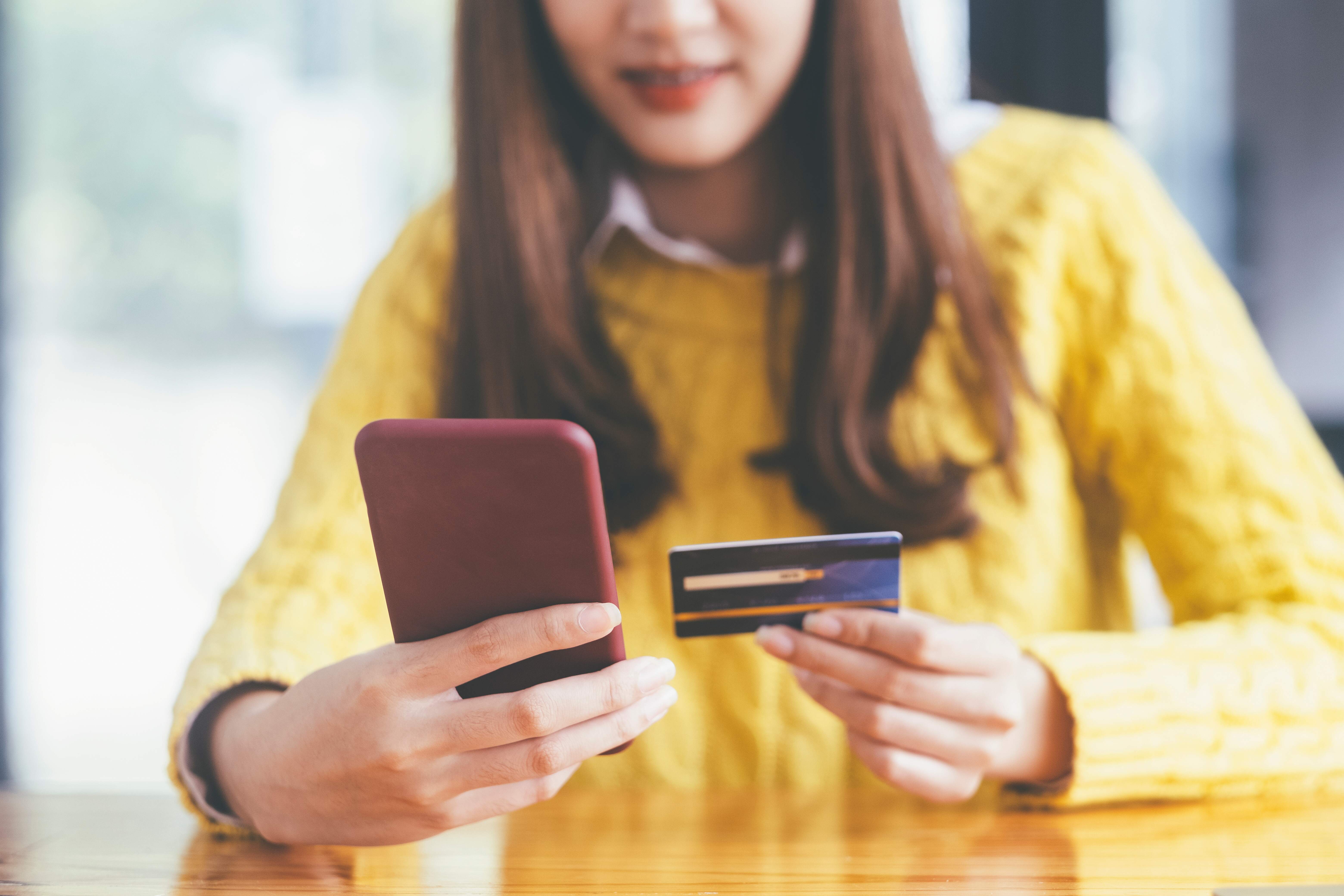 Mulher branca, segurando um cartão de crédito e olhando o celular.