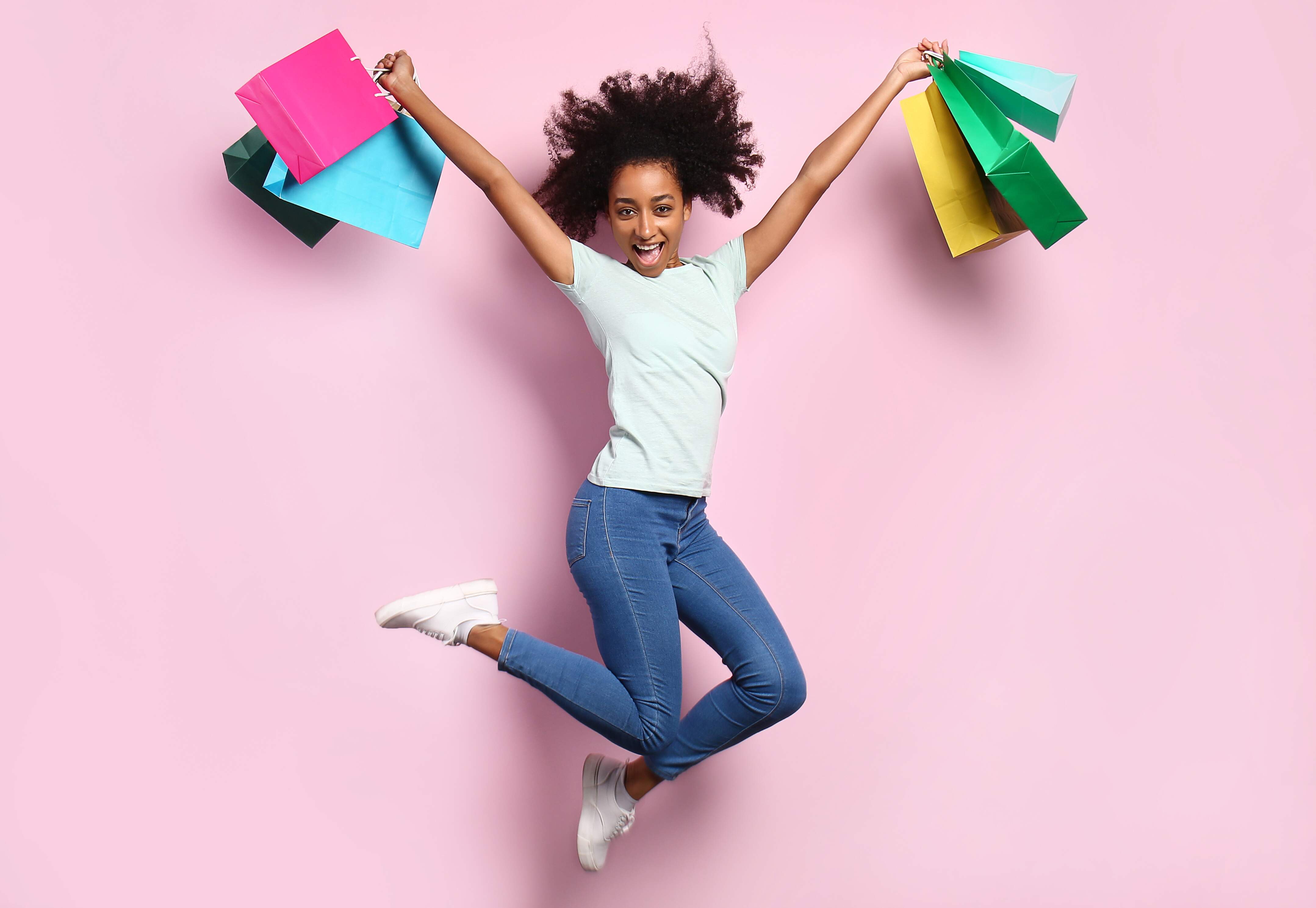 Mulher negra saltando feliz com sacolas de compras coloridas nas duas mãos em um fundo rosa