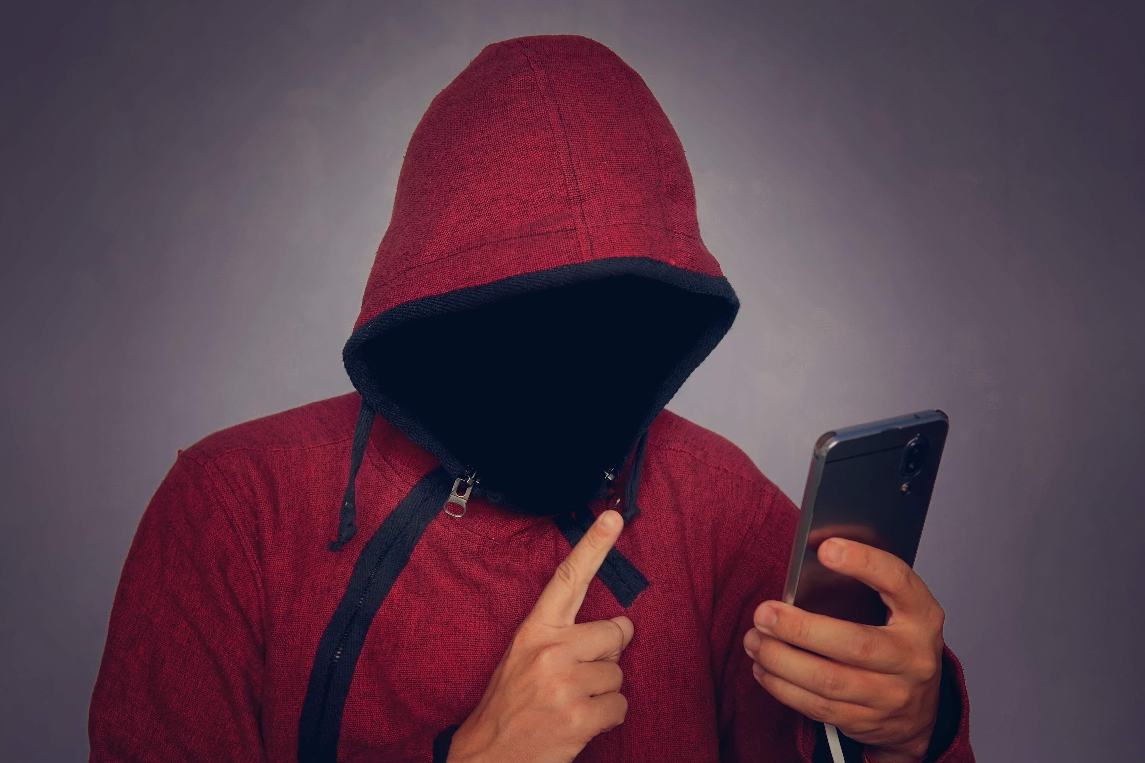 Pessoa com um moleton de capuz e ziper, com o rosto oculto dentro do capuz, segurando um celular, com um fundo escuro atrás.