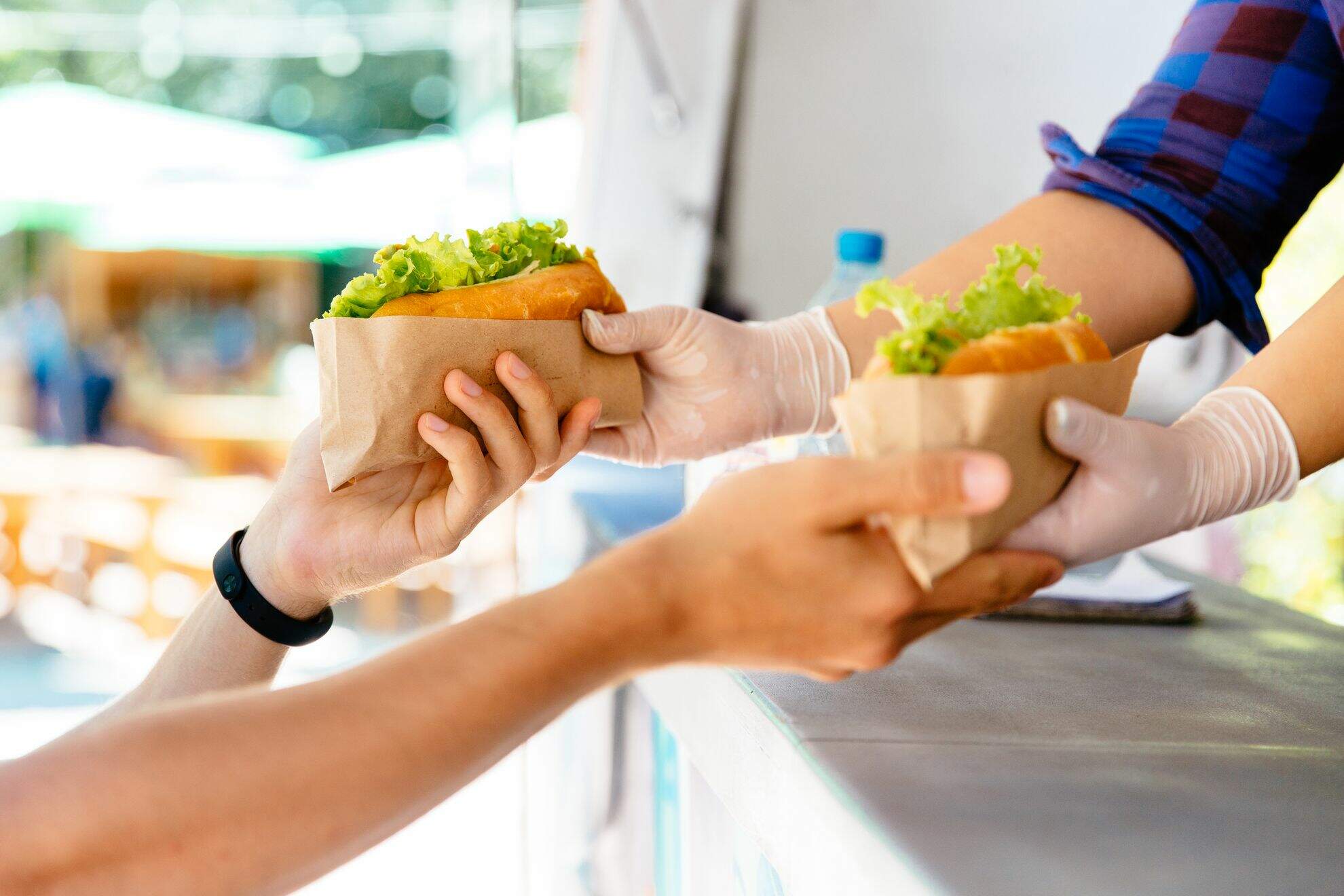 Mãos saindo de dentro de um foodtruck, entregando sanduíches para uma pessoa.